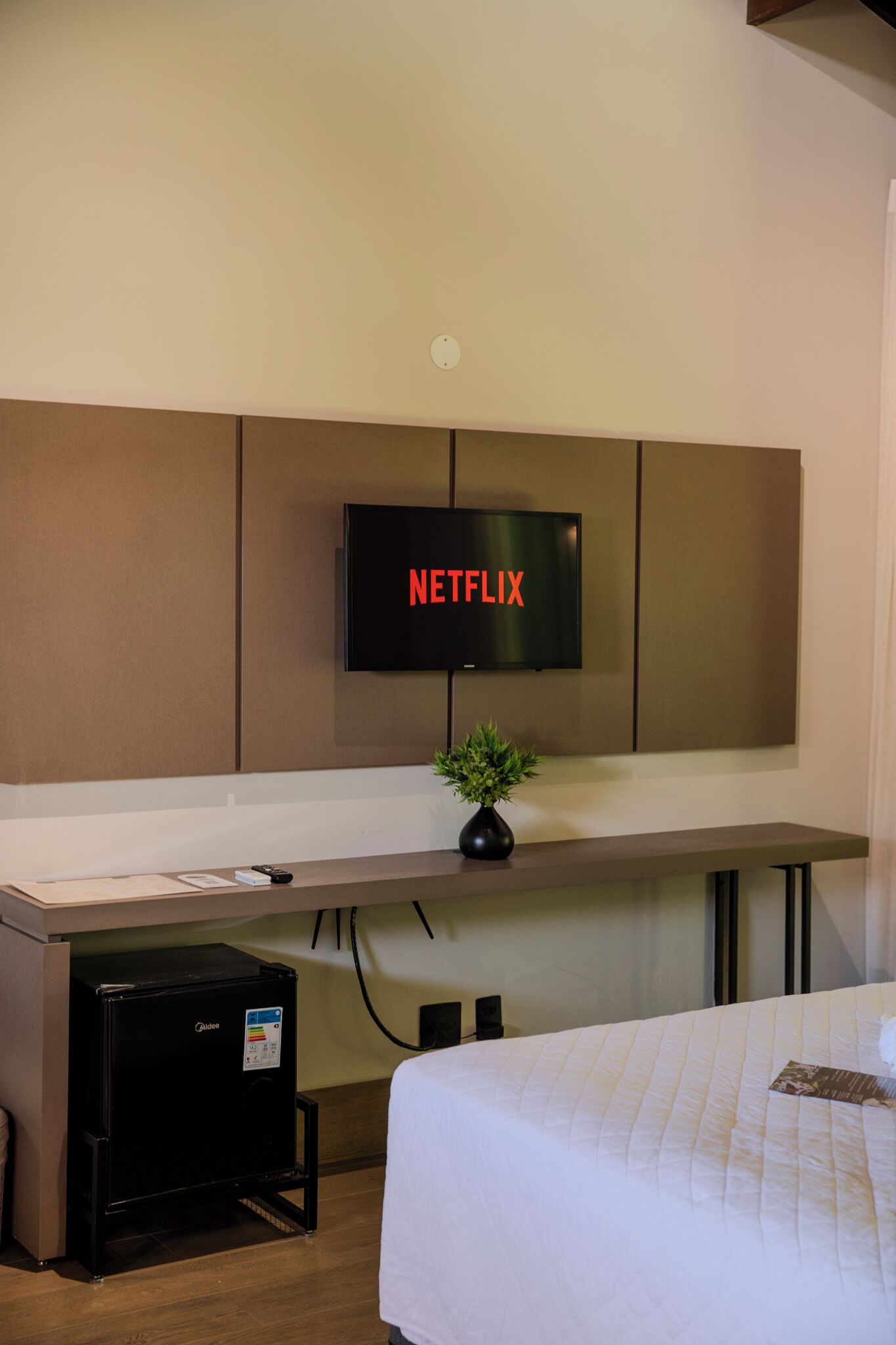 A hotel room with a bed, a desk with a computer, a small refrigerator, a TV mounted on the wall, and a plant on the desk.