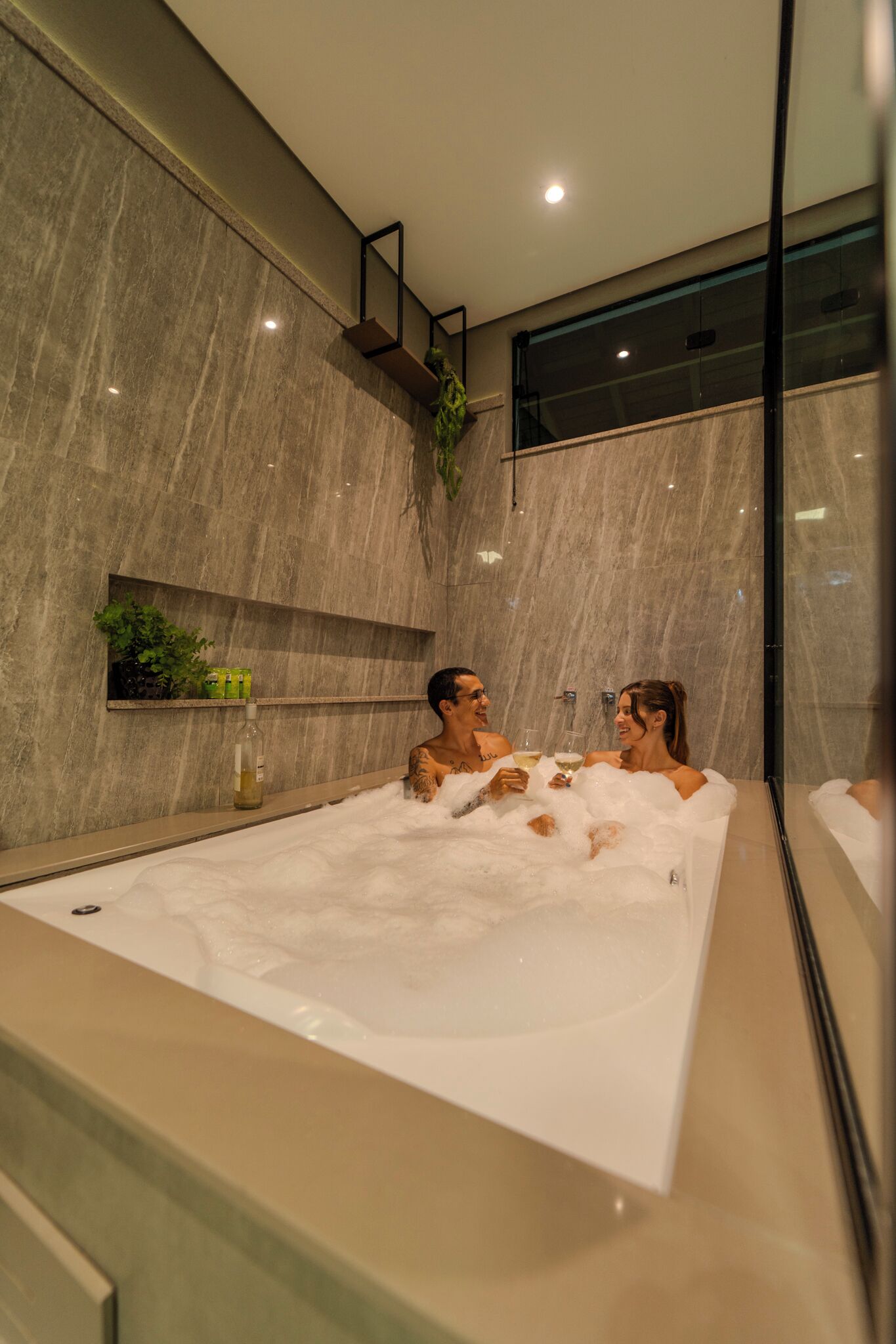 A modern bathroom with a large bathtub, marble walls, and a plant on a shelf.