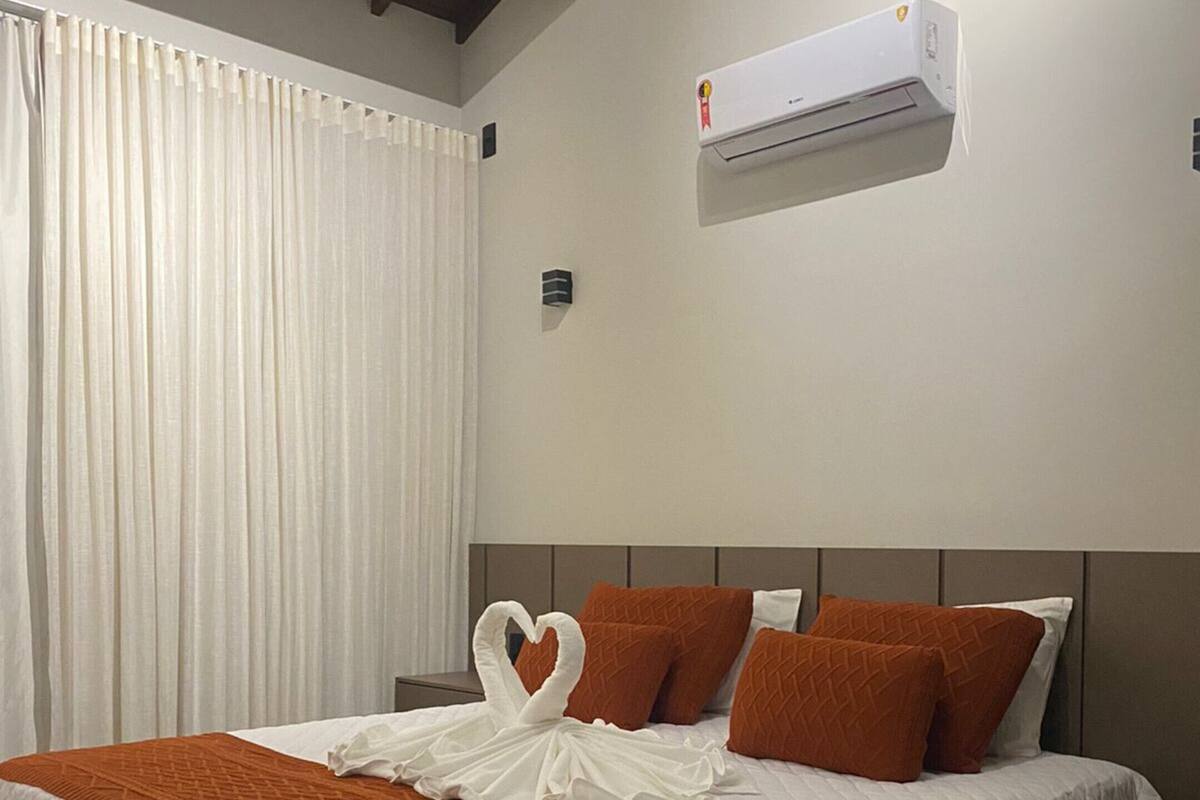 Una habitación de hotel con una cama, cortinas blancas, un equipo de aire acondicionado y almohadas naranjas.