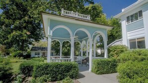 Een overdekt terras met witte zuilen en een witte balustrade, omringd door groen en een pad naar een huis.