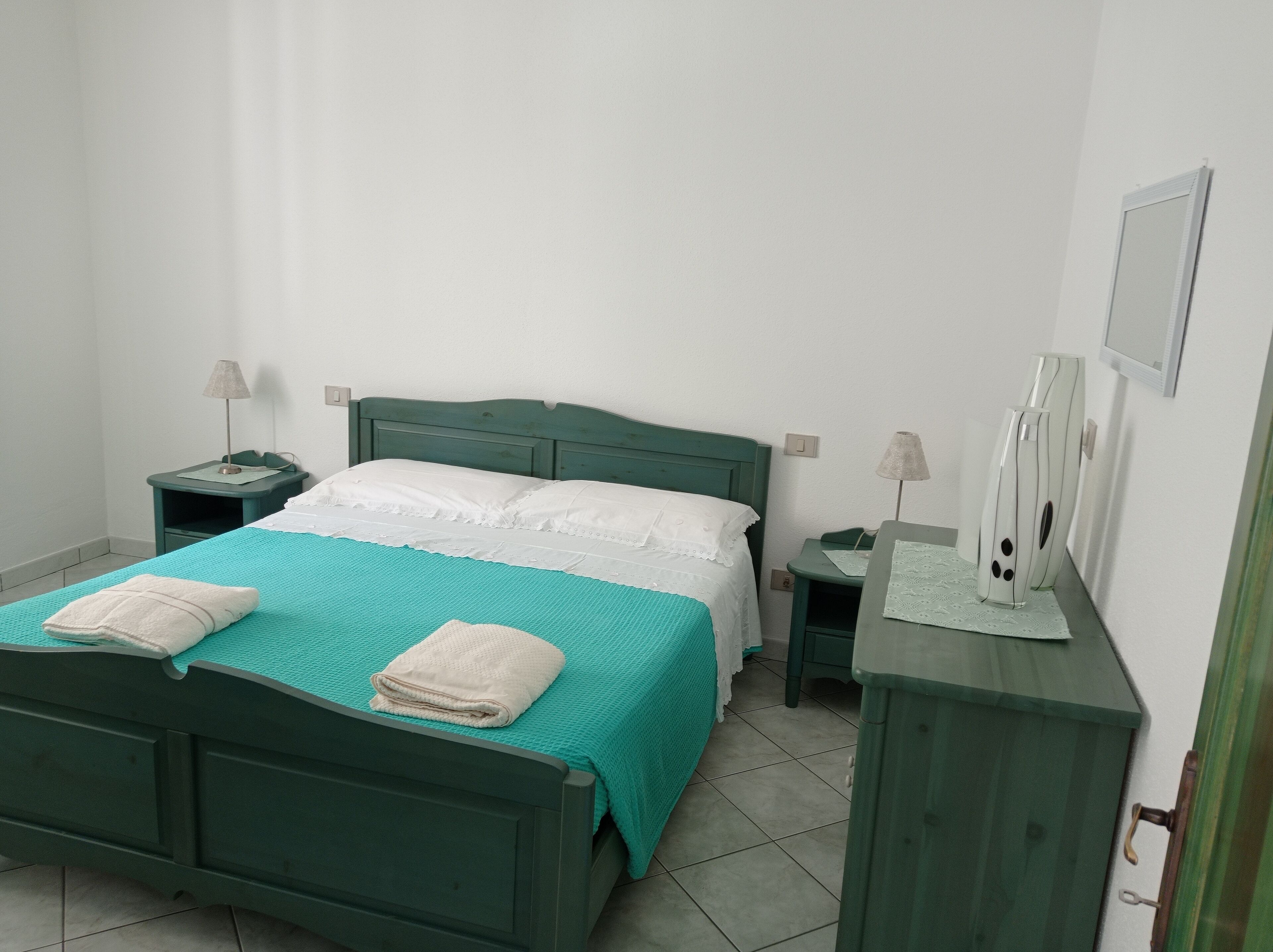 Un lit bien fait, recouvert d’une couvre-lit turquoise et agrémenté d’oreillers blancs, une petite table de chevet avec une lampe, et une table de nuit également dotée d’une lampe.