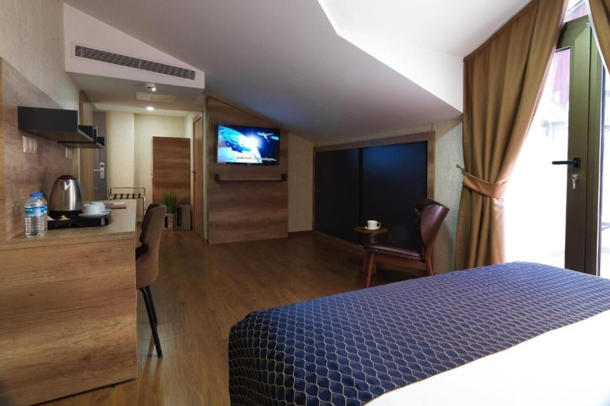A hotel room with a bed, a desk with a kettle, a chair, a television, and a window with curtains.
