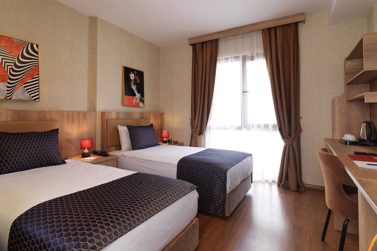 A hotel room with two beds, a desk, and a chair. There is a window with curtains, a painting on the wall, and a nightstand with a lamp.