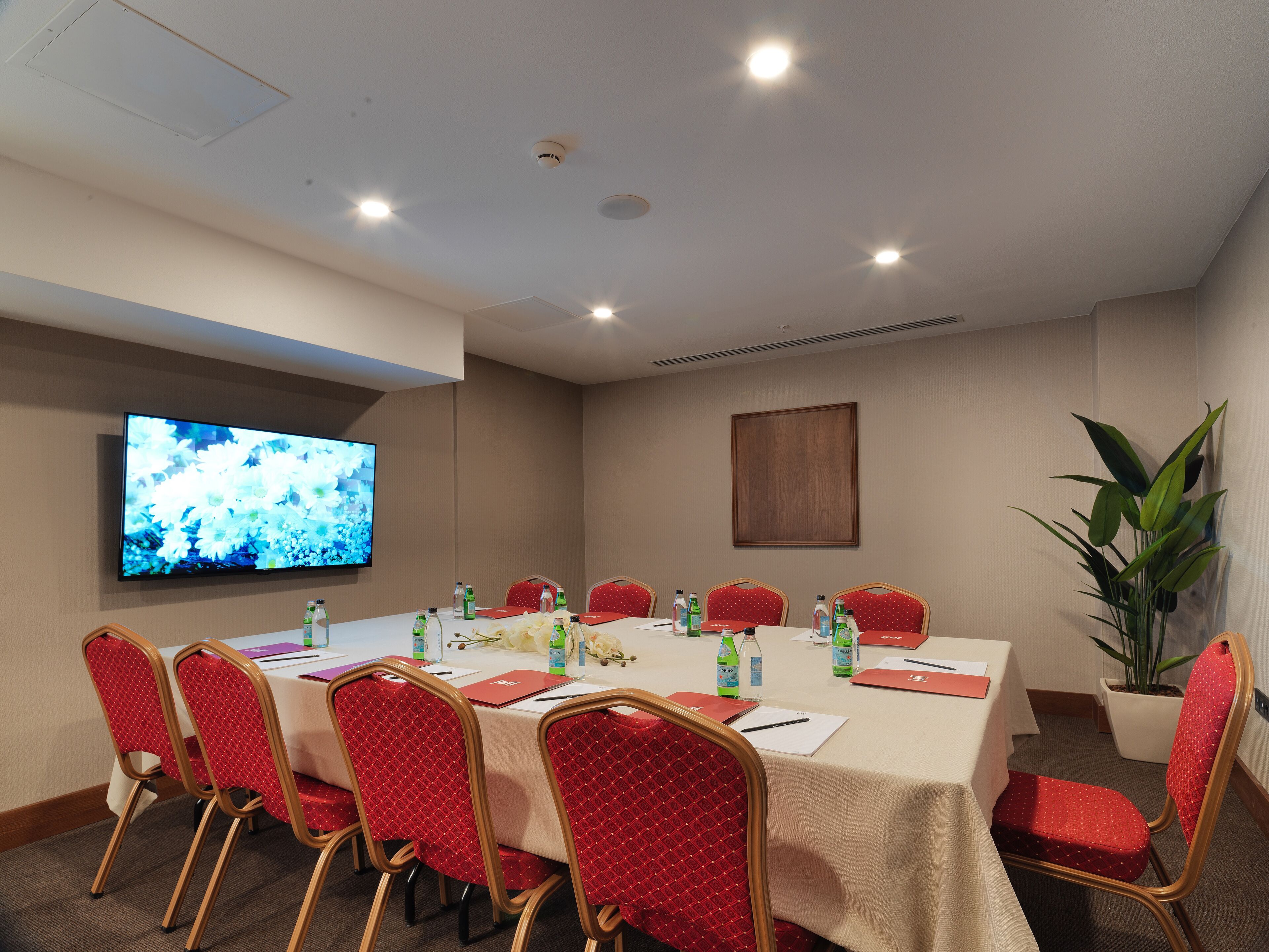 A conference room with a large flat-screen TV, red upholstered chairs, a white table with name cards, water bottles, and a potted plant.