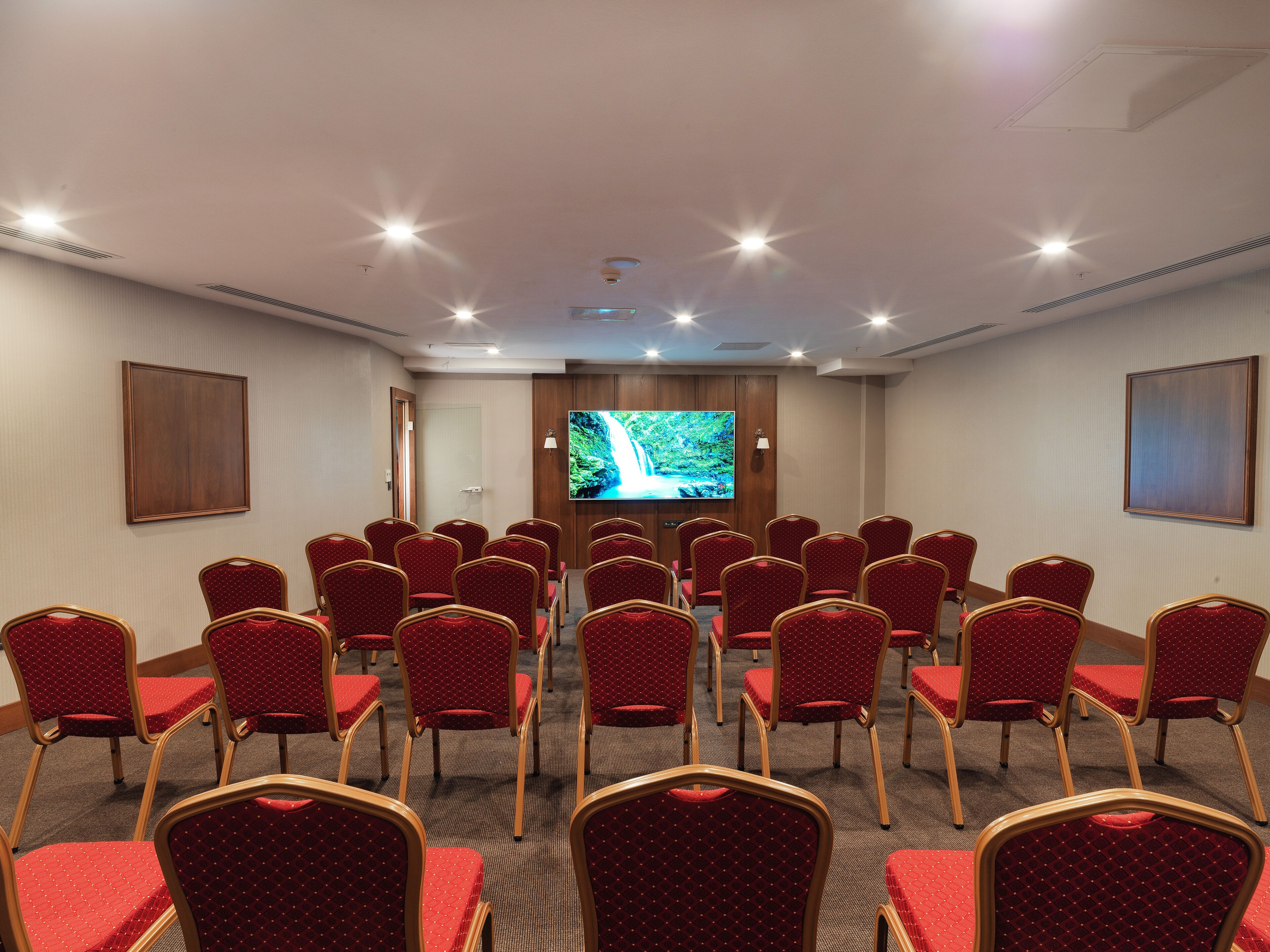 A conference room with rows of red chairs, a large screen, and a door in the background.