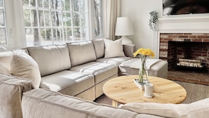 Un salon avec un canapé d’angle beige, une table basse ronde en bois sur laquelle est posé un vase de fleurs jaunes, une lampe blanche et une cheminée.
