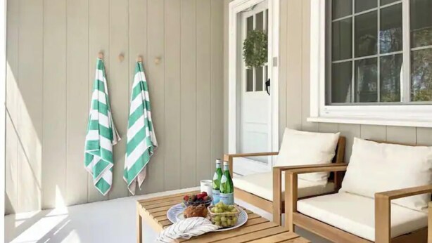Une véranda baignée de soleil, avec une table en bois dressée pour un repas, des fruits et des boissons, un fauteuil confortable et des serviettes à rayures accrochées au mur.