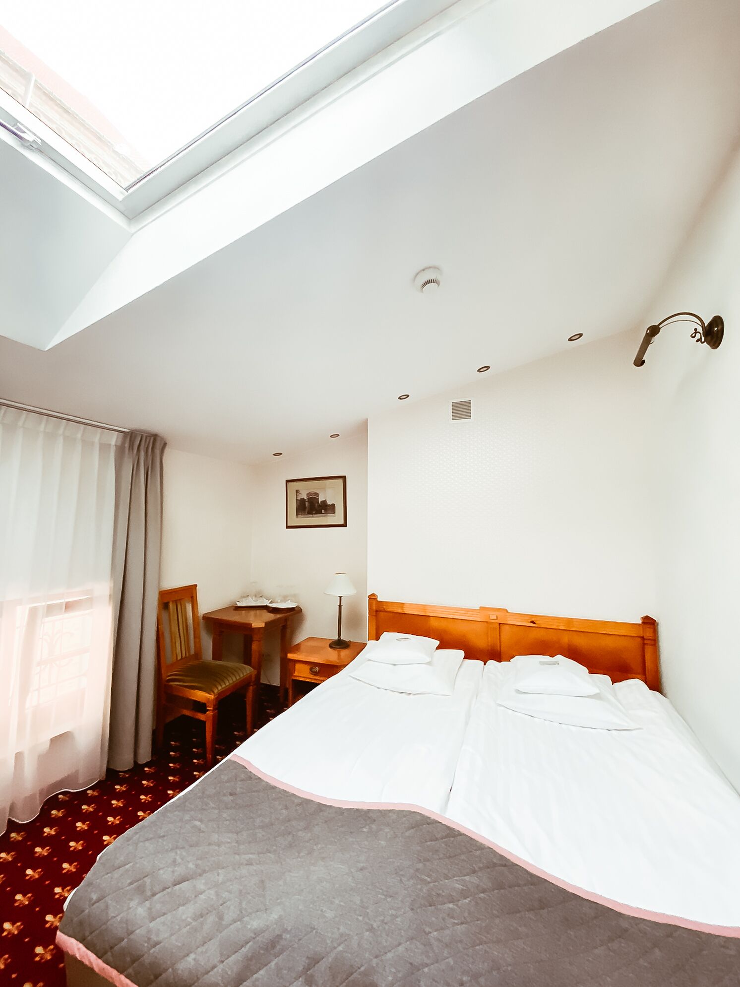 Habitación de hotel con tragaluz, una cama, un escritorio, una silla y un cuadro en la pared.