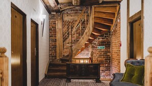Una escalera con barandillas de madera que baja a un nivel inferior, un banco y un cojín verde en una habitación con paredes de ladrillo y puertas de madera.