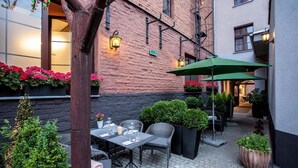 Un patio al aire libre con mesas y sillas, rodeado de macetas con plantas y un muro de ladrillo.