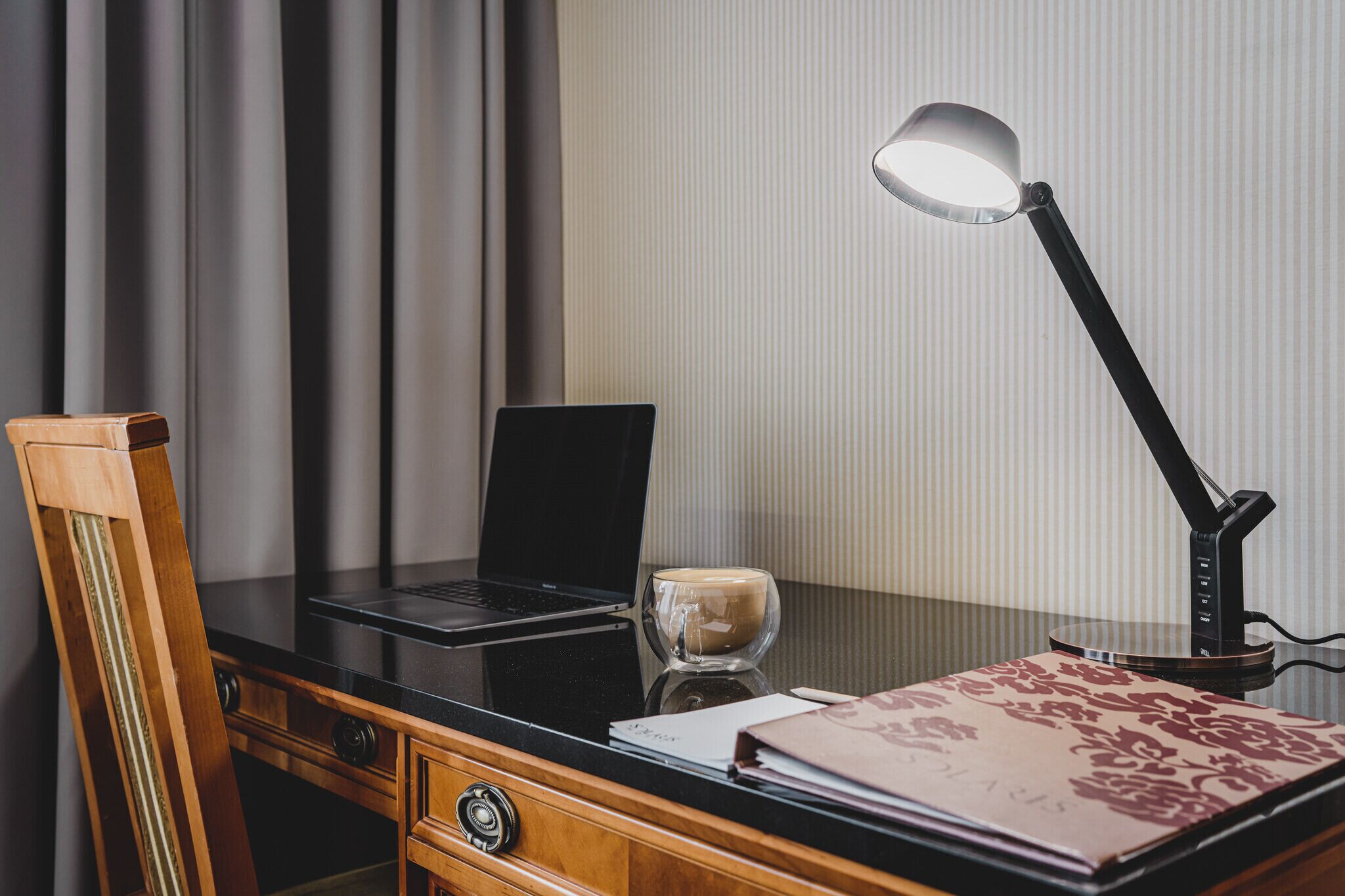 A wooden desk with a laptop, a glass coffee cup, a lamp, and a patterned folder.