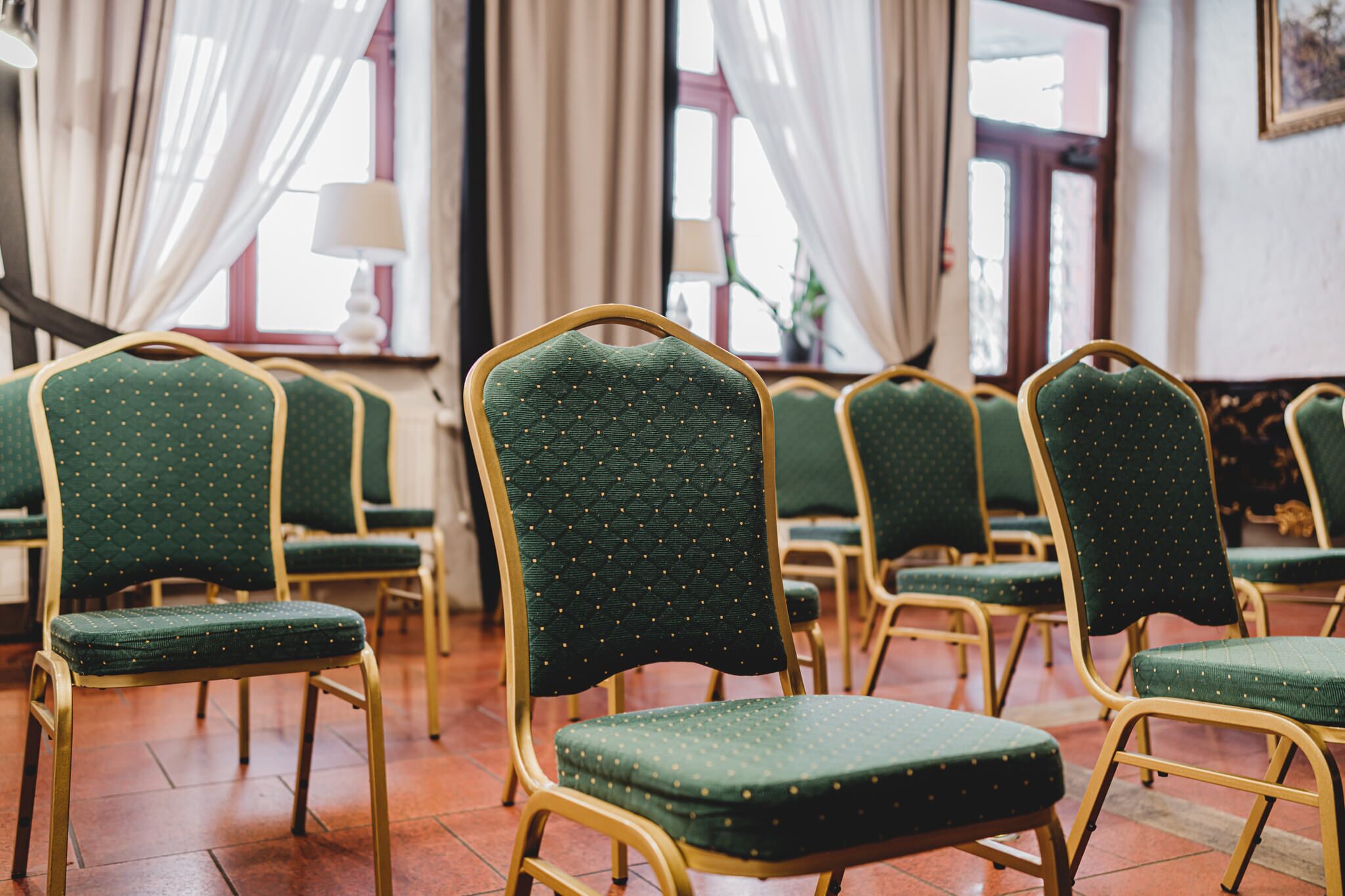 De nombreuses rangées de fauteuils verts, recouverts de tissu et dotés de cadres dorés, sont disposées dans une pièce aux grandes fenêtres et aux rideaux.