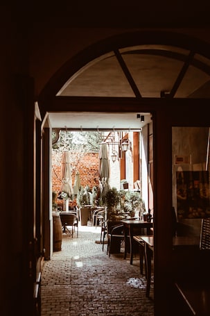 A cobblestone pathway leading to an outdoor seating area with tables and chairs, a brick wall, and hanging umbrellas.