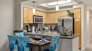 A modern kitchen with wooden cabinets, stainless steel appliances, and blue chairs. - Ko'Olina Oasis: Lagoon, Pool & Resort Living (Kapolei)