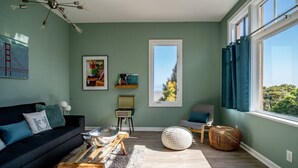 Una sala de estar moderna con un sofá negro, un taburete blanco redondo, una mesa de centro y un ventanal con cortinas.