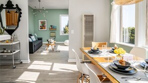 Un comedor con una mesa larga dispuesta para cuatro, una zona de cocina con un carrito y una sala de estar con un sofá y un cuadro.