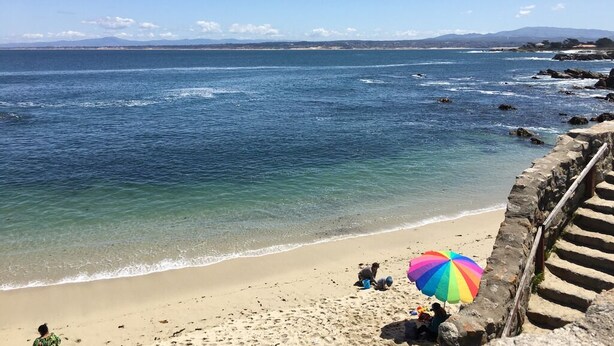 Una playa con agua azul cristalina, orilla arenosa y un paraguas colorido.