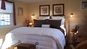 A neatly made bed with white bedding, two framed floral pictures on the wall, a bedside lamp, and a wicker chair.