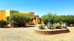 A building with a yellow facade, surrounded by greenery and a gravel courtyard.