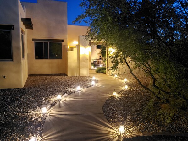 A pathway lined with lights leading to a building entrance, surrounded by gravel and trees.
