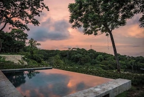 Uma piscina infinita retangular, com borda transparente, cercada por vegetação exuberante e árvores, refletindo o céu.