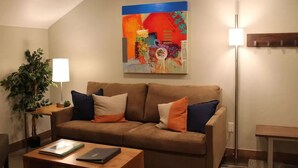 Un salon avec un canapé marron, deux coussins, une table basse sur laquelle se trouvent un livre et un ordinateur portable, une lampe sur pied, un tableau accroché au mur et une plante en pot.