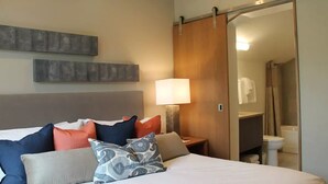 Une chambre d’hôtel comprenant un lit, des oreillers, une table de chevet avec une lampe, une porte coulissante donnant accès à la salle de bain et une œuvre d’art accrochée au mur.