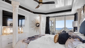 A bedroom with a large bed, a ceiling fan, a sunburst mirror, and a view of the ocean.