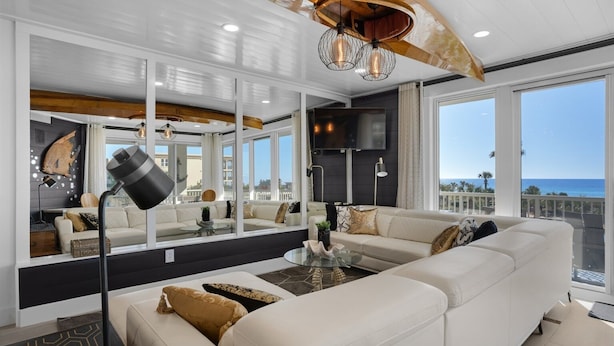 A modern living room with a large sectional sofa, a coffee table, and a floor lamp. There is a large mirror reflecting the room and a television mounted on the wall.