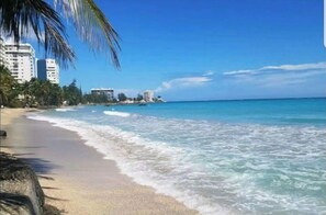 Una playa con aguas azul cristalino, una orilla arenosa y edificios al fondo.