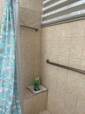 Zona de ducha con paredes de azulejos, cortina de baño con estampado de conchas marinas y un estante con artículos de aseo.