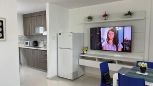 Eine moderne Küche mit integriertem Mikrowellenherd, Kaffeemaschine und einem weißen Kühlschrank. Es gibt einen Essbereich mit Tisch und blauen Stühlen, sowie einen an der Wand montierten Flachbildfernseher, der eine Szene aus einer Fernsehsendung zeigt.