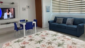Ein modernes Wohnzimmer mit einem blauen Sofa, einem Esstisch mit blauen Stühlen, einem an der Wand befestigten Fernseher und einem großen Fenster mit Jalousien.