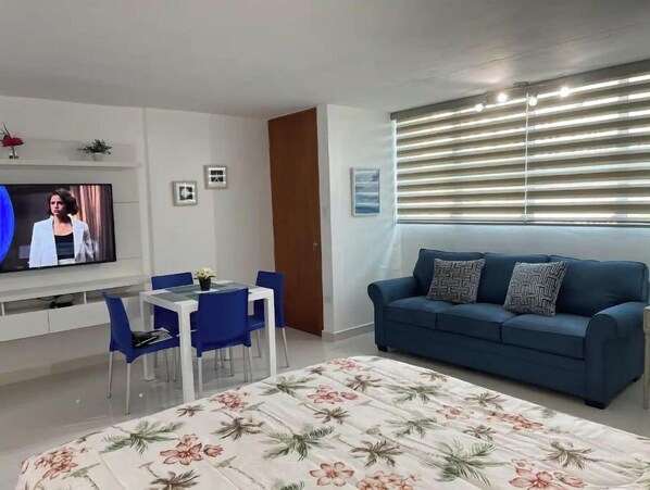 Una sala de estar moderna con un sofá azul, una mesa de comedor con sillas azules, un televisor montado en la pared y un ventanal con persianas.