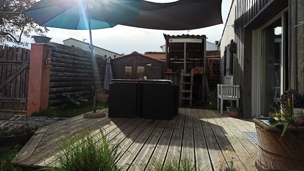 Una terraza de madera con zona sombreada, cerca de madera y un edificio con un cartel que dice 'PLAGE'.