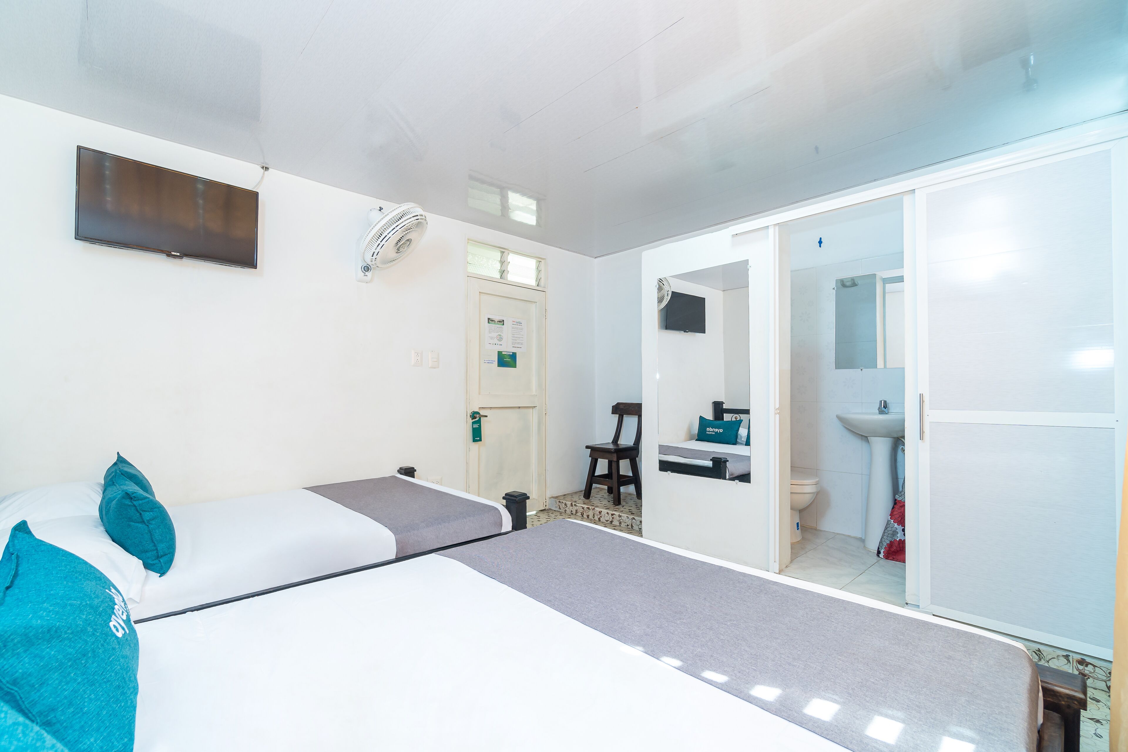Quarto com cama, escrivaninha, cadeira, televisão, ventilador e banheiro visível através de uma porta aberta.