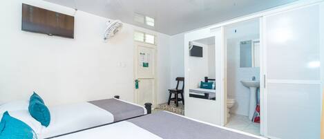 Quarto com cama, escrivaninha, cadeira, televisão, ventilador e banheiro visível através de uma porta aberta.
