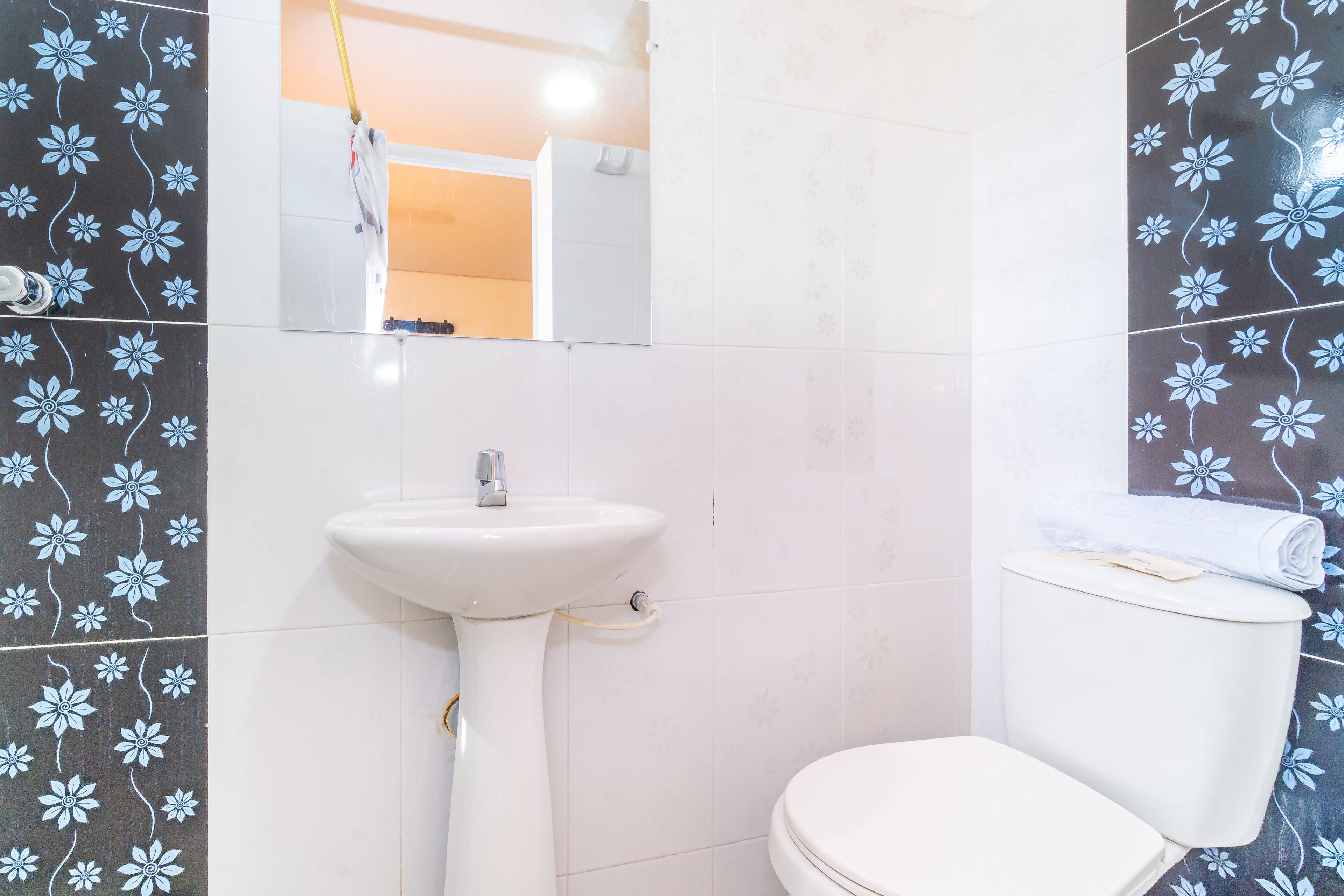 Un baño con lavamanos, inodoro y espejo blancos. Las paredes tienen un estampado de flores.