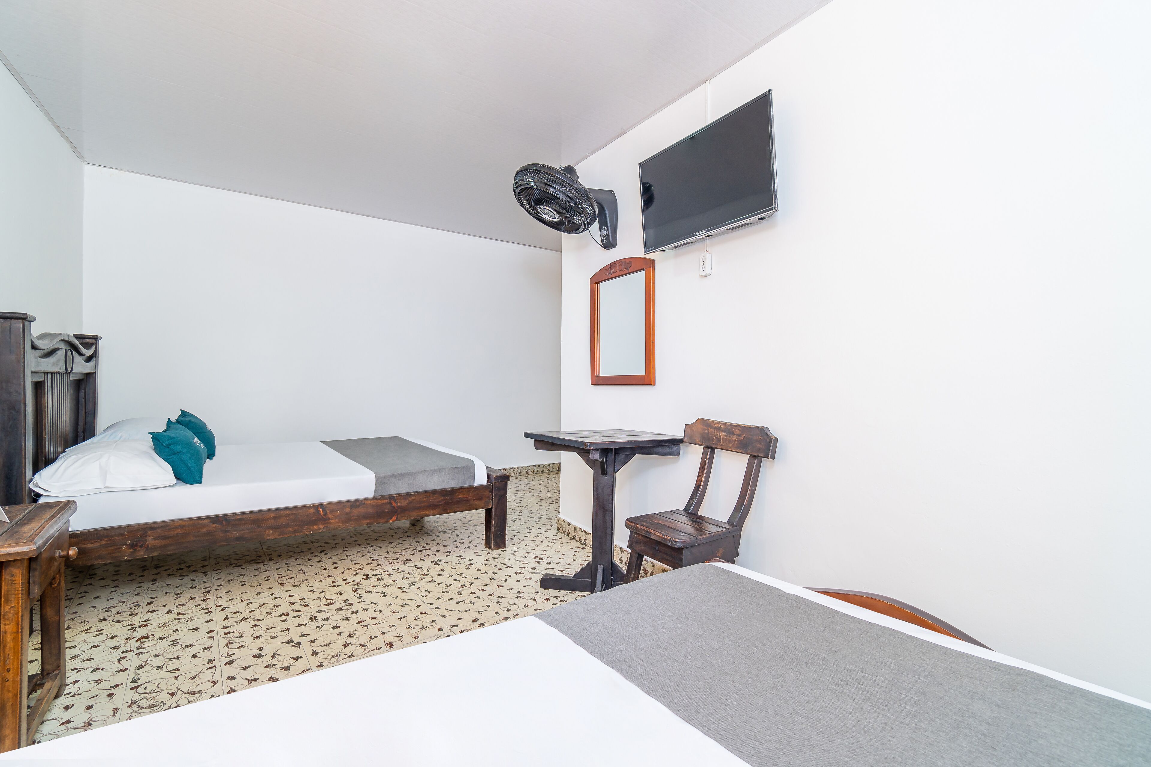 A compact room with a bed, a small table, a chair, a wall-mounted TV, and a ceiling fan.