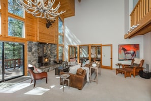 A spacious living room with a stone fireplace, wooden ceiling, and large windows.