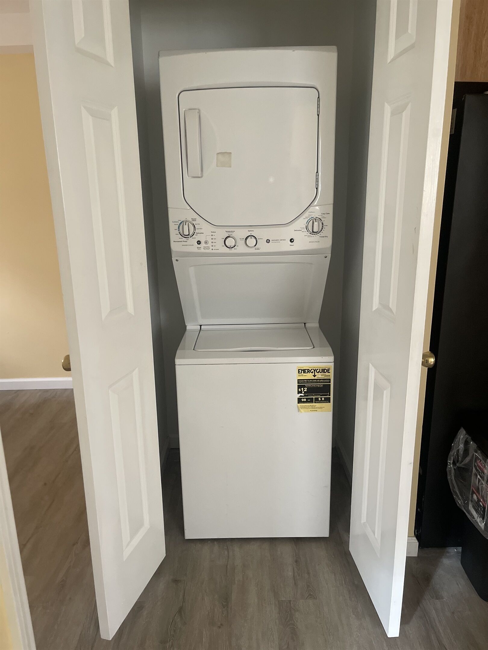 A compact, white front-loading washer and dryer unit in a narrow space with a wooden floor, a white door on the left, and a black door on the right.