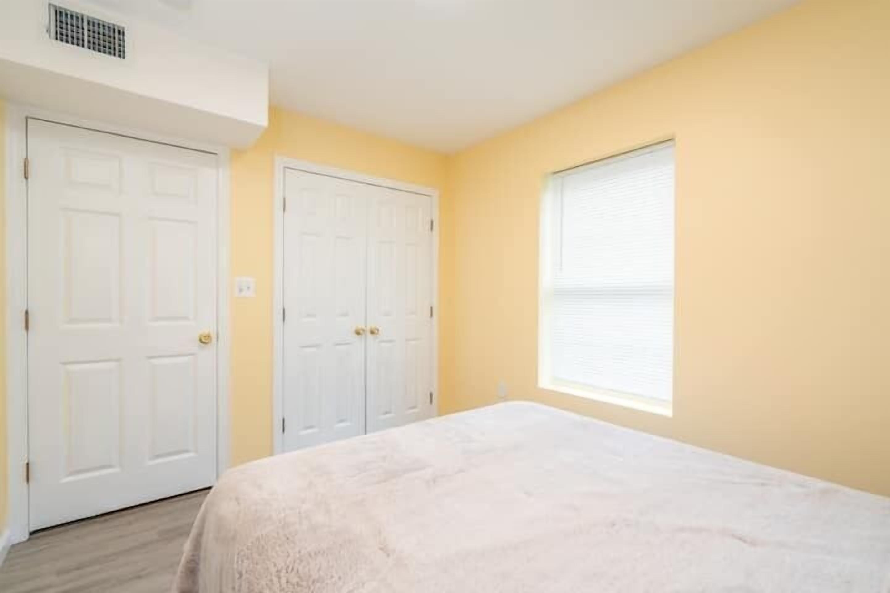 Quarto com duas portas brancas, uma janela com persianas e uma cama com cobertura branca.