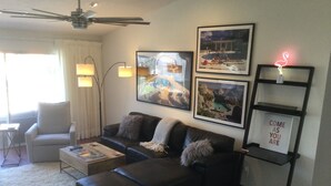 A modern living room with a black leather sofa, a white fluffy rug, and a ceiling fan. There are framed pictures on the wall and a neon flamingo sign on a shelf.