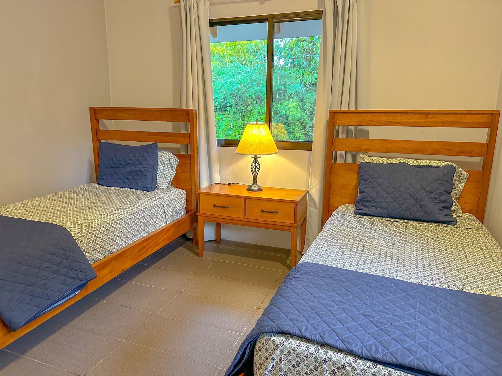 A room with two single beds, each with a quilted blue bedspread and a matching pillow. There is a wooden nightstand between the beds with a lamp and a drawer. A window with curtains is visible, showing greenery outside.