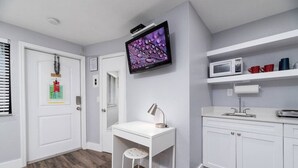 A modern, well-lit room with a white desk, a mounted TV, a microwave, and a small shelf with cups. - Shell on the Beach #104 (Hollywood)