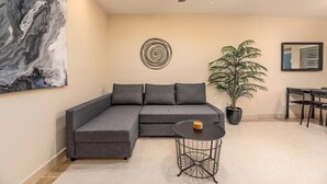 Un salon moderne avec un canapé d’angle gris, une table ronde en métal, une plante en pot et des œuvres d’art accrochées aux murs.