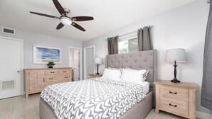 Quarto com cama, duas mesas de cabeceira, ventoinha no teto, janela com cortinas e porta.
