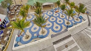Un hôtel situé en bord de mer, avec une cour dallée aux motifs géométriques, des palmiers et une jetée en arrière-plan.