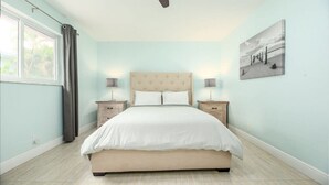 Une chambre à coucher avec un grand lit, deux tables de chevet, un ventilateur de plafond, une fenêtre avec des rideaux et un tableau accroché au mur.
