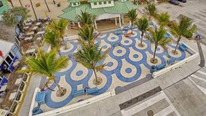 Ein Strandhotel mit Innenhof, der Palmen und ein gemustertes Pflaster besitzt, einer in den Ozean führenden Steg und einem Parkplatz mit mehreren Autos.